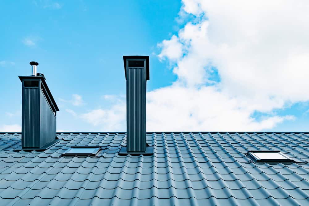 A close-up view of a modern metal rooftop with a blue, shiny finish. The roof features two prominent chimneys, hinting at recent Chimney Leak Repairs Twin Cities MN, and two small skylights. The background showcases a clear, blue sky with a few scattered clouds.