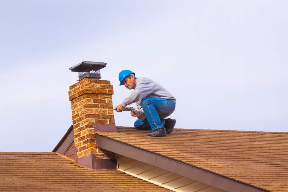 A person wearing a blue hard hat and casual clothing repairs a brick chimney on the rooftop of a building. They are using a tool to work on the mortar. The sky is clear behind them.