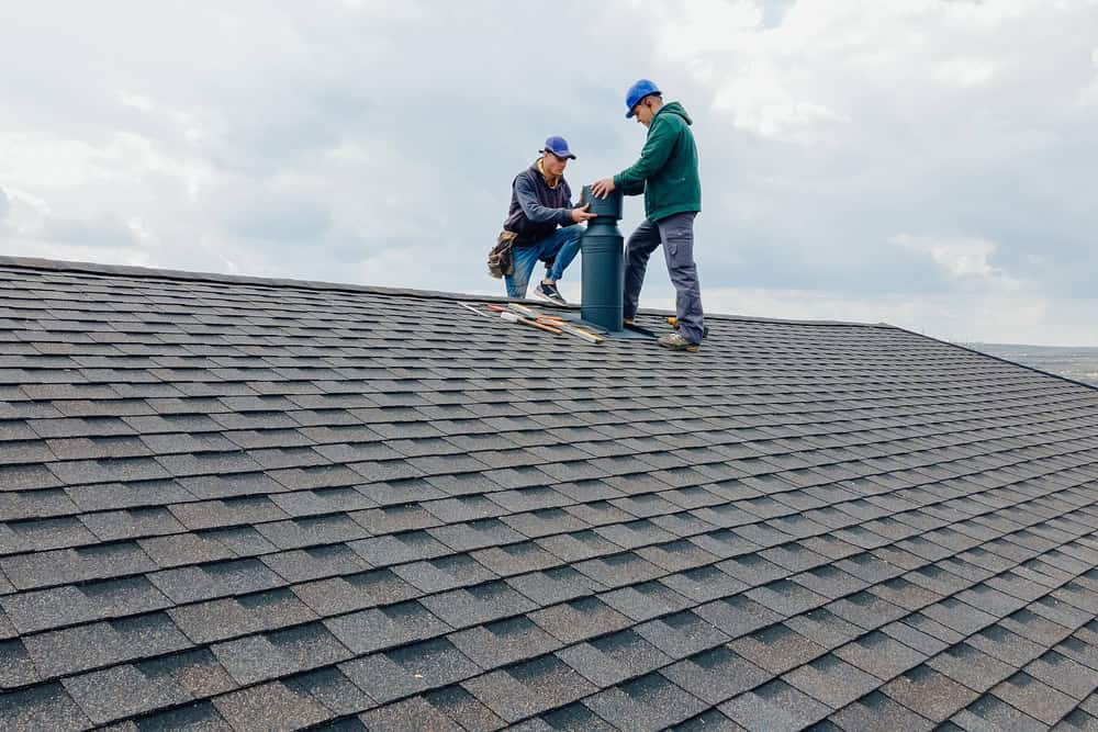 Two workers in safety gear and blue caps are repairing a rooftop vent on a shingled roof under a cloudy sky. They have tools and materials spread around them.