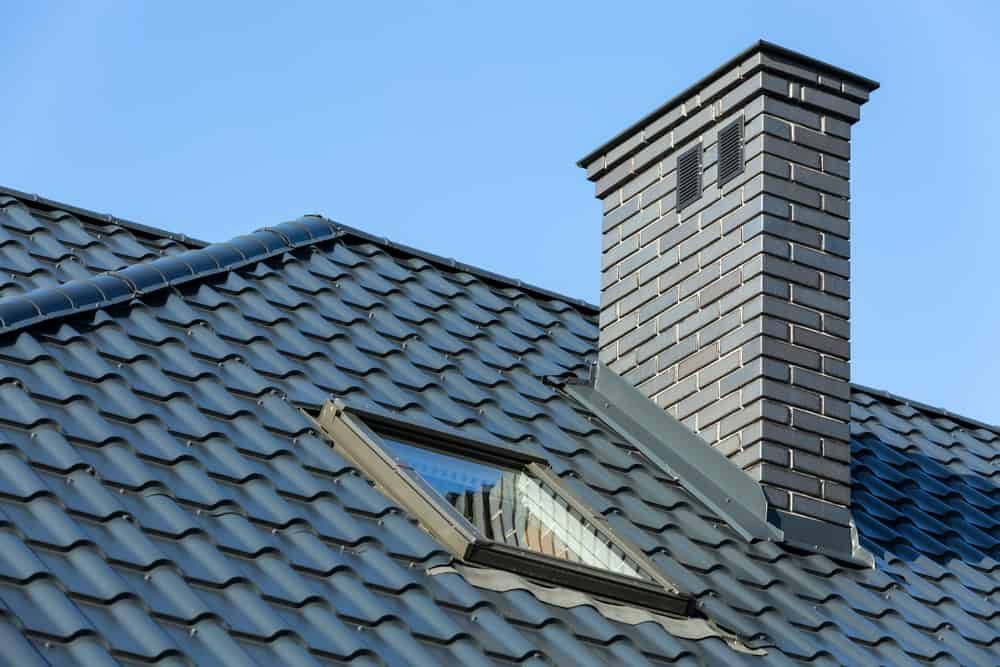 Close-up of a house roof featuring blue, wavy metal tiles. A rectangular window is partially open in the roof, and a gray brick chimney stands prominently. The sky in the background is clear and blue.