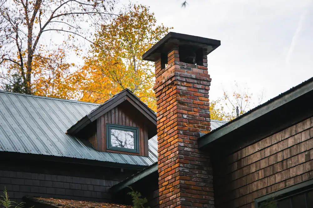 A close-up of a brick chimney on a wooden house with a metal roof, surrounded by trees with autumn-colored leaves in the background.