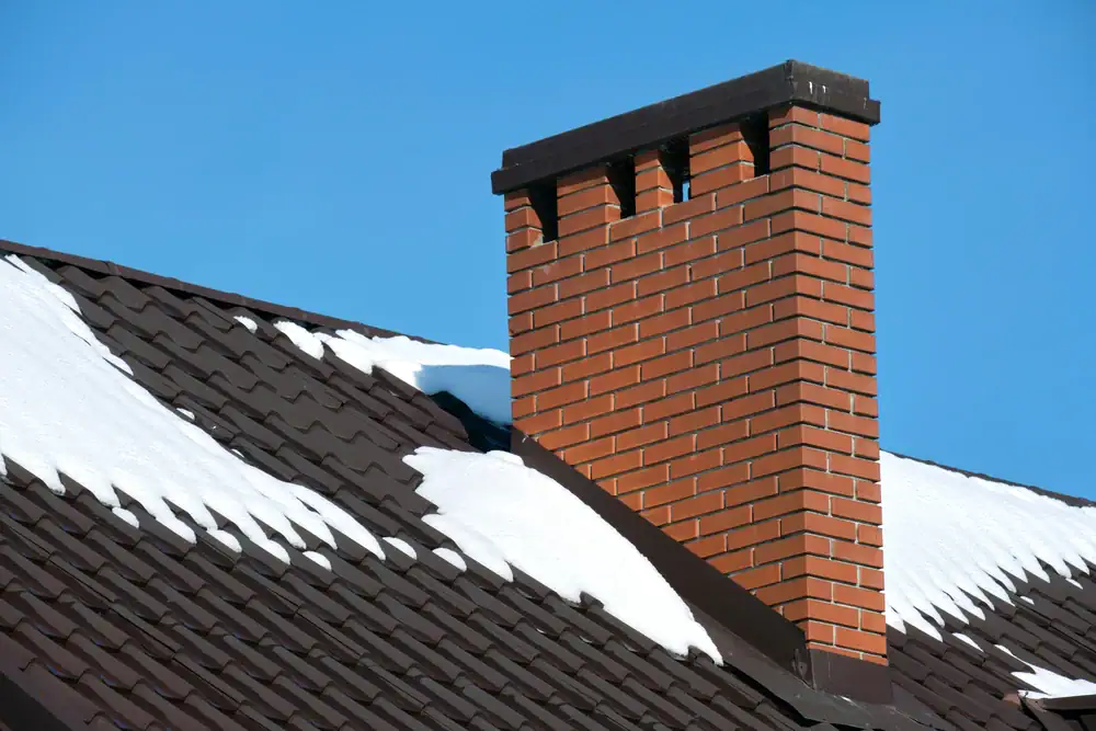 A red brick chimney rises from a dark brown tiled roof, partially covered with patches of melting snow, set against a clear blue sky.