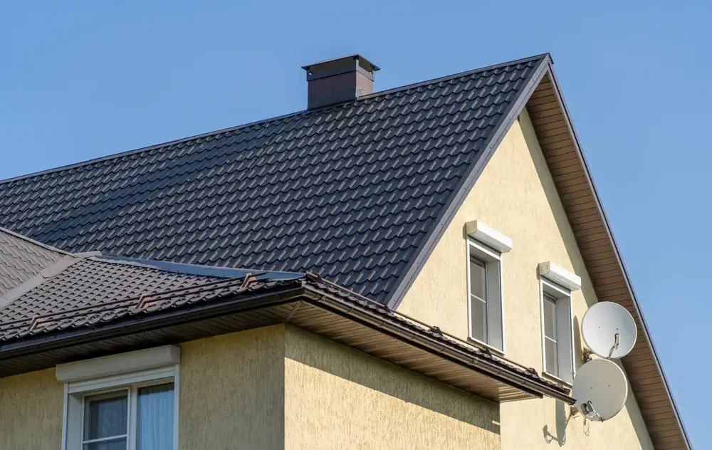 The image shows the upper part of a house with a sloped, dark metal roof, beige walls, white-framed windows, a chimney, and two satellite dishes mounted on the side, all under a clear blue sky.