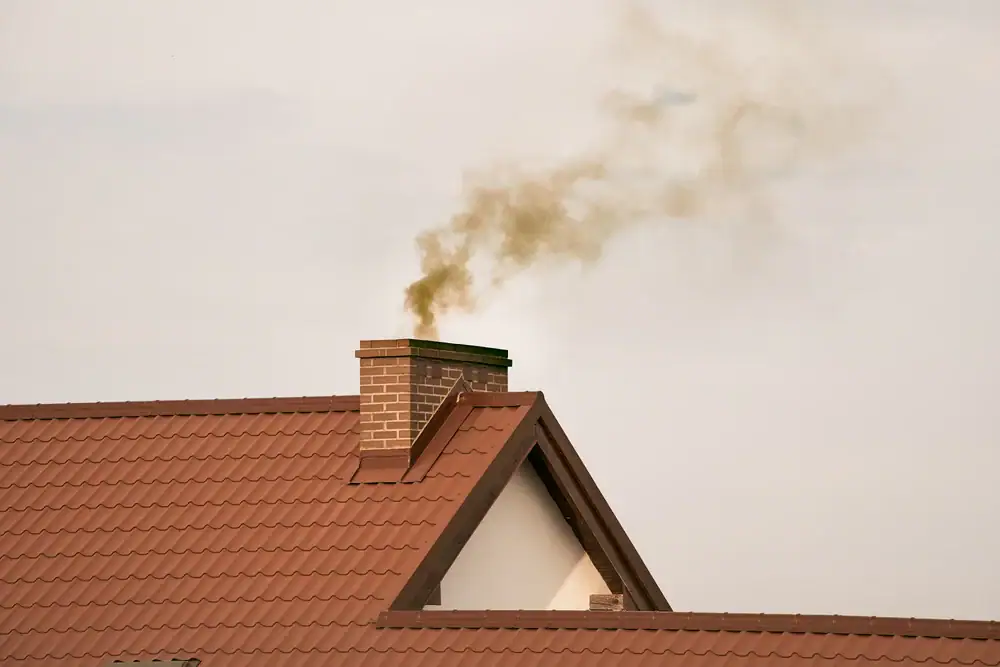 A brown brick chimney on a red-tiled roof emits a plume of grayish smoke into a pale, overcast sky.