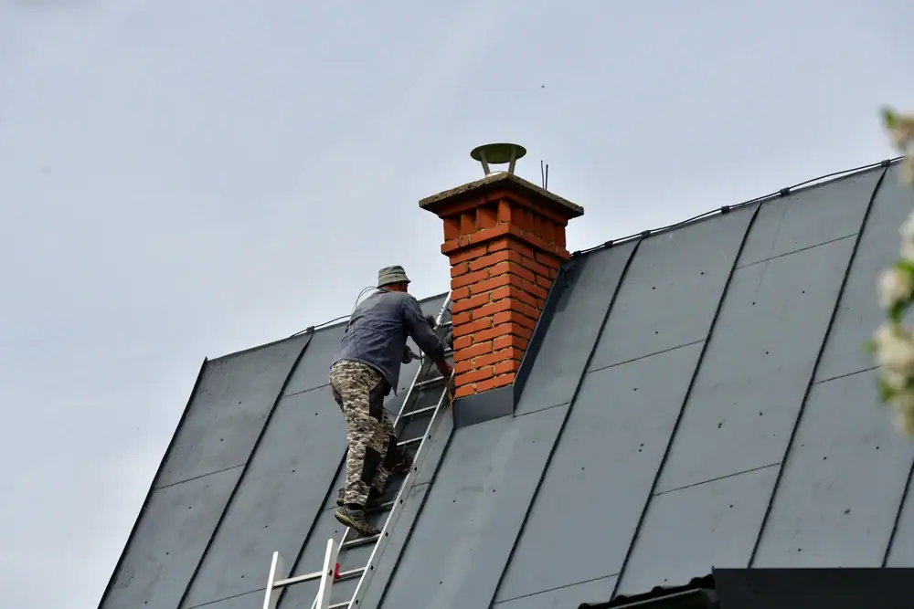 A person in camouflage pants and a hat stands on a ladder leaning against a steep metal roof, inspecting or working on a red brick chimney under a cloudy sky.